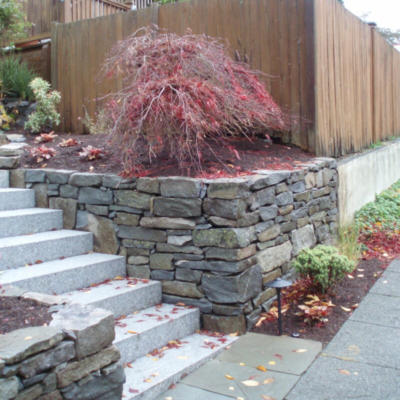 Rock retaining wall seen on either side of large granite stairwell leads to the front door of residential home in Seattle, Washington.