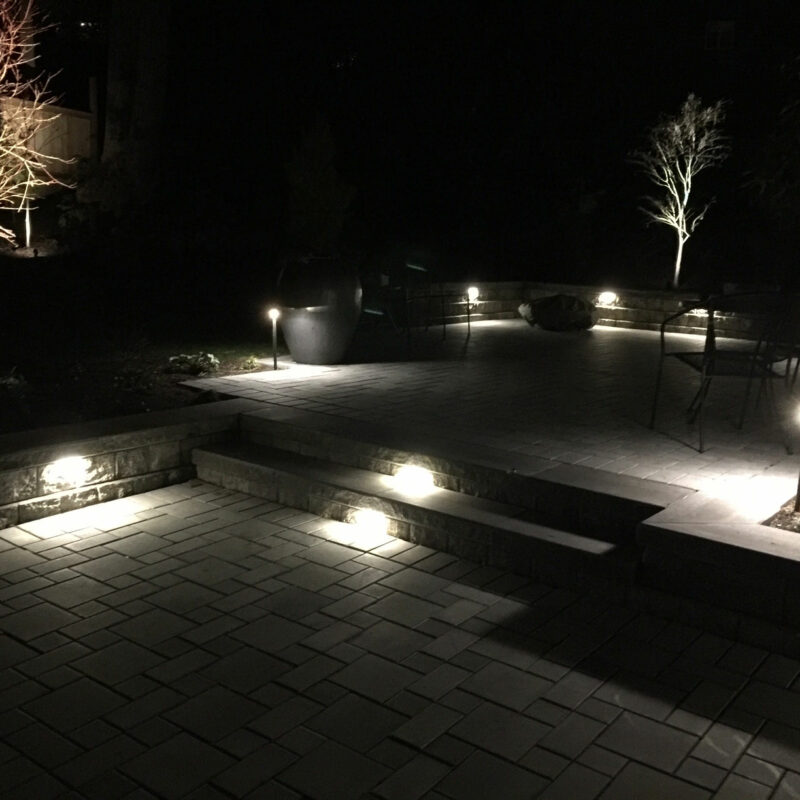 Backyard paver patio seen at night is illuminated with custom outdoor lighting from Bonney Lake Landscapes located in Seattle, Washington.
